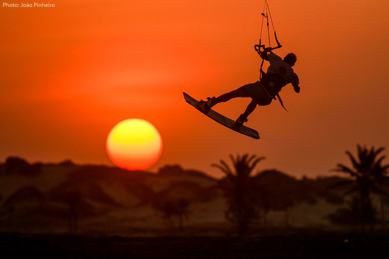 http://Silhouette%20d'un%20kitesurfeur%20effectuant%20un%20saut%20aérien%20en%20contre-jour%20devant%20un%20grand%20soleil%20couchant%20orange%20au-dessus%20des%20dunes%20et%20palmiers%20au%20Brésil.