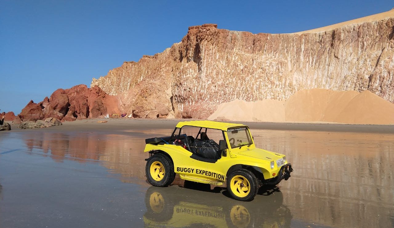 A dune buggy sits on the shimering sand with an ocher cliff in the background. 
