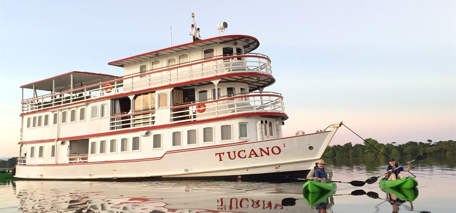 Vue du bateau de croisière de charme en Amazonie Tucano sur l'eau dans le soleil couchant avec 2 kayakistes devant.