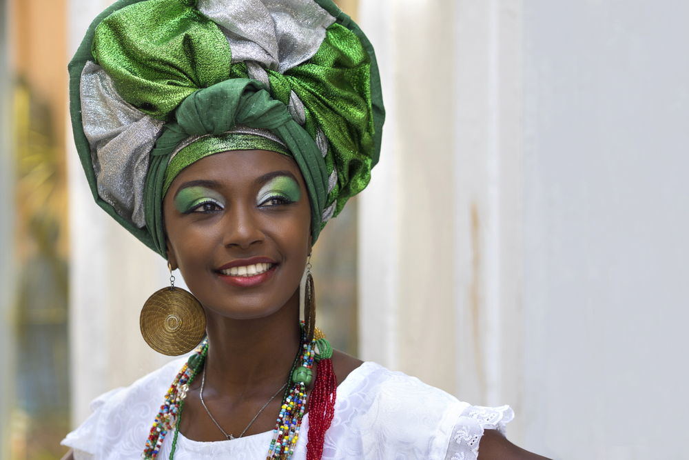 http://Portrait%20rapproché%20d'une%20femme%20afro-brésilienne%20souriante%20à%20Salvador,%20Brésil.%20Elle%20porte%20une%20coiffe%20traditionnelle%20complexe%20appelée%20torso%20en%20vert%20et%20argent,%20ainsi%20qu'un%20maquillage%20vert%20scintillant%20sur%20ses%20paupières.%20De%20grandes%20boucles%20d'oreilles%20dorées%20et%20plusieurs%20colliers%20de%20perles%20colorées%20ornent%20son%20cou.%20Elle%20porte%20un%20haut%20blanc%20à%20volants.%20L'image%20capture%20la%20beauté,%20la%20fierté%20et%20l'héritage%20culturel%20de%20la%20région%20de%20Bahia,%20en%20mettant%20en%20valeur%20les%20couleurs%20vibrantes%20et%20les%20textures%20de%20ses%20vêtements%20et%20accessoires.%20L'arrière-plan%20est%20flou,%20mais%20laisse%20entrevoir%20l'architecture%20coloniale%20de%20Salvador.