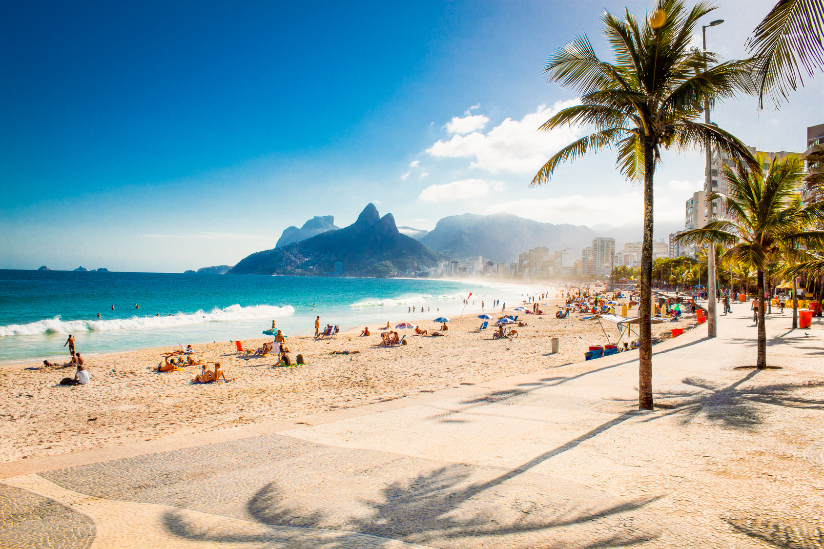 http://Vue%20panoramique%20de%20la%20plage%20d'Ipanema%20à%20Rio%20de%20Janeiro,%20Brésil,%20par%20une%20journée%20ensoleillée.%20La%20photo%20est%20prise%20depuis%20la%20promenade%20en%20bord%20de%20mer,%20montrant%20le%20sable%20doré,%20les%20eaux%20turquoise%20de%20l'océan%20Atlantique%20et%20les%20montagnes%20iconiques%20Dois%20Irmãos%20en%20arrière-plan.%20Des%20baigneurs%20et%20des%20amateurs%20de%20soleil%20sont%20visibles%20sur%20la%20plage.%20Un%20palmier%20se%20dresse%20au%20premier%20plan,%20projetant%20son%20ombre%20sur%20la%20promenade.%20L'atmosphère%20est%20détendue%20et%20invitante,%20invitant%20à%20la%20détente%20et%20à%20la%20découverte%20de%20la%20nature.