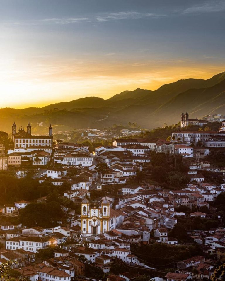 Vue panoramique en plongée de la ville coloniale d'Ouro Preto au Brésil lors du coucher du soleil, avec ses maisons blanches aux toits rouges, ses églises baroques et les montagnes du Minas Gerais en arrière-plan baignées d'une lumière dorée.