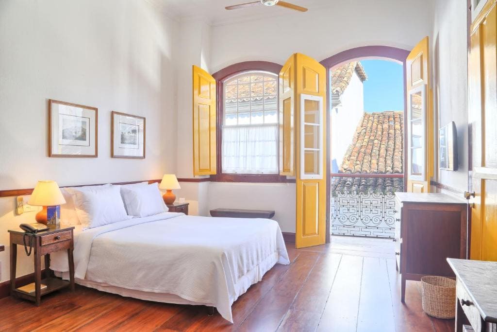 Chambre de style colonial à la Pousada do Ouro em Paraty, avec lit double blanc, parquet en bois et balcon avec vue sur les toits historiques.