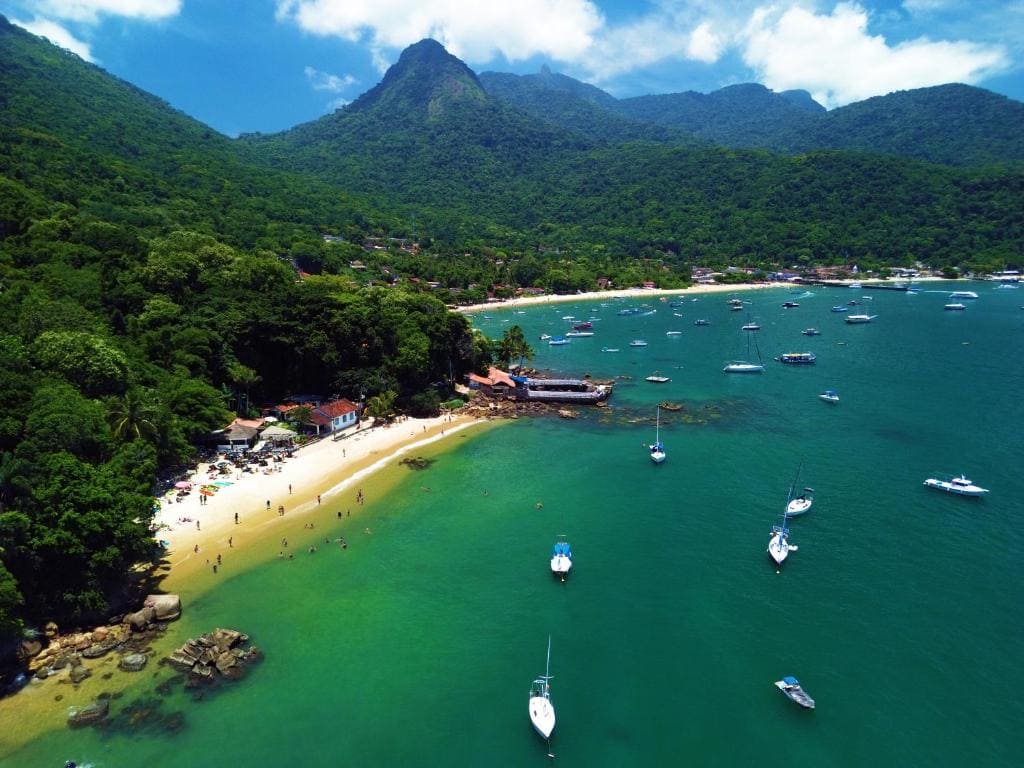 Vue aérienne de la baie d'Abraão à Ilha Grande avec ses eaux turquoise, ses nombreux bateaux, sa plage de sable fin et ses montagnes verdoyantes en arrière-plan.