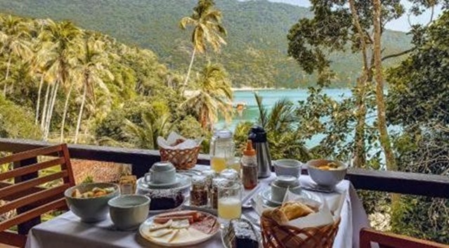 http://Table%20de%20petit-déjeuner%20garnie%20de%20fruits,%20jus,%20pains%20et%20cafés%20sur%20une%20terrasse%20en%20hauteur%20offrant%20une%20vue%20sur%20les%20cocotiers,%20la%20forêt%20tropicale%20et%20la%20mer%20turquoise%20d'Ilha%20Grande%20au%20Brésil.