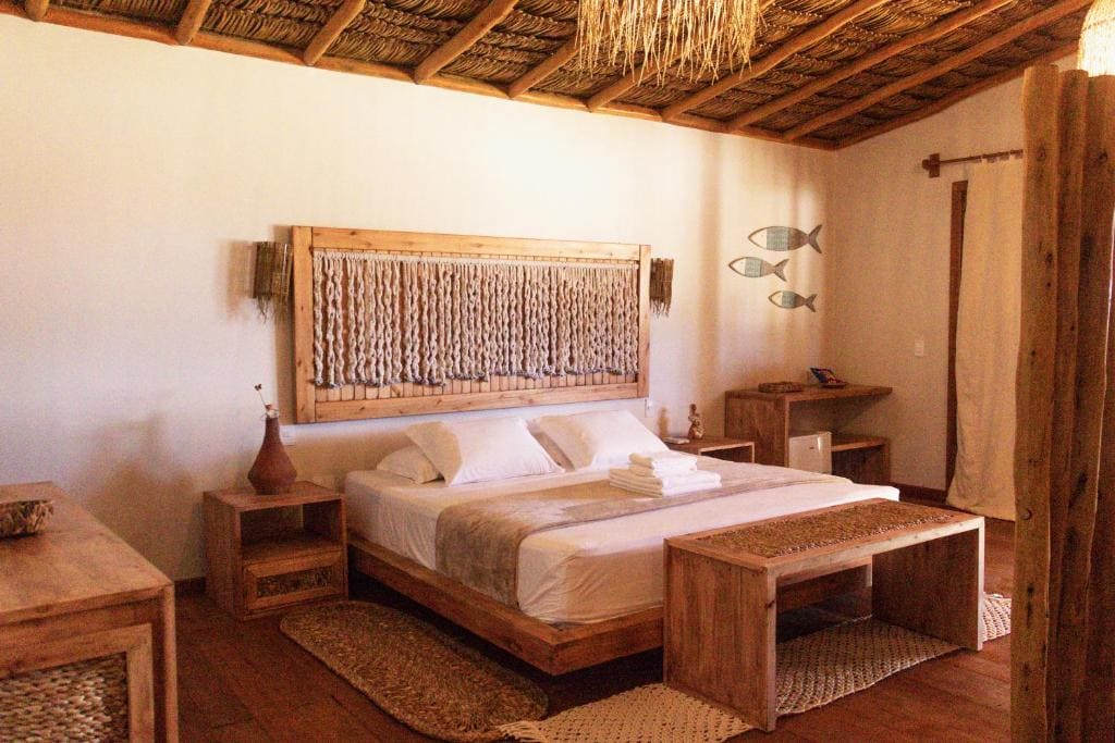 Vue latérale oblique d'une chambre d'hôtel de style pousada, montrant un lit avec une tête de lit en macramé unique, un plafond en chaume, des poissons sculptés sur le mur de droite, et un grand pilier en bois dans le premier plan.