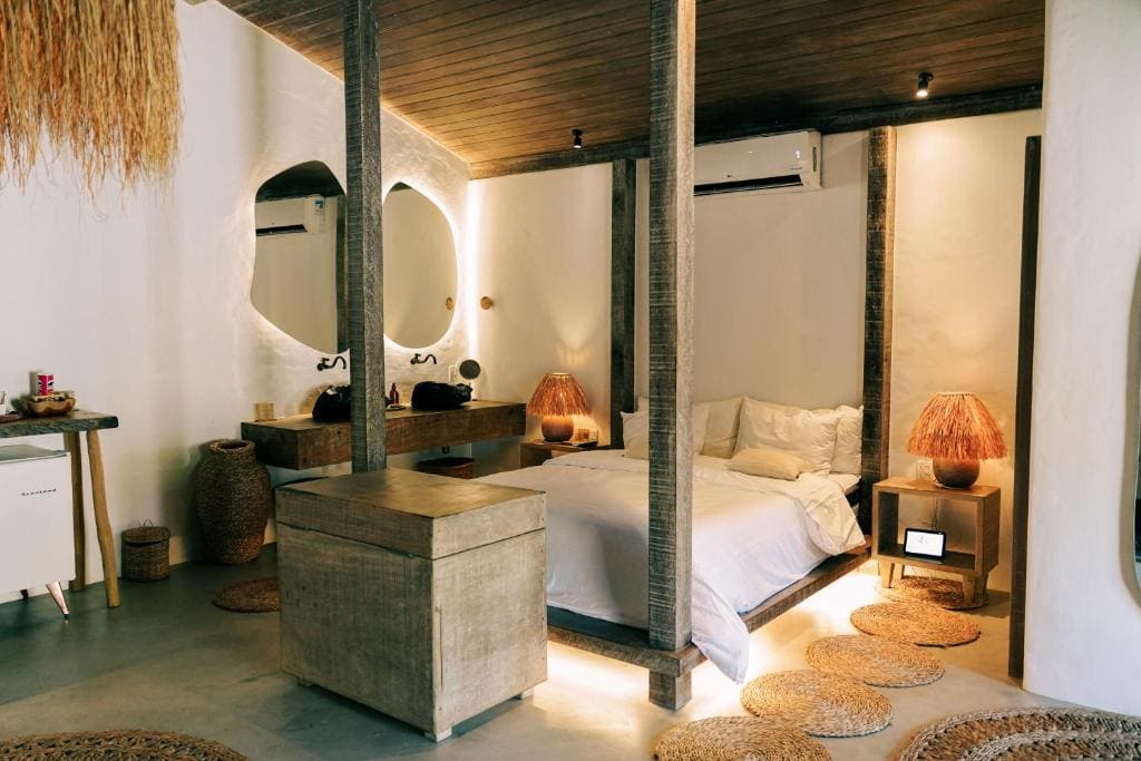Vue intérieure d'une chambre élégante à l'Anacardier Pousada à Atins, mettant en avant un lit à colonnes en bois, un décor aux matières naturelles et une ambiance lumineuse chaleureuse sur un sol en béton poli.