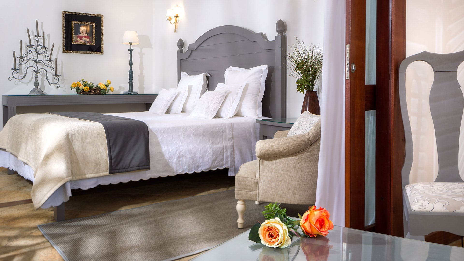 Intérieur d'une chambre d'hôtel de luxe. Un grand lit central avec tête de lit grise est dressé avec du linge blanc et des jetés gris. Les tables de chevet en verre encadrent le lit. Au fond, une console avec des fleurs et un chandelier est sous un tableau encadré. Au premier plan, une table basse en verre montre deux roses. Un fauteuil beige est à gauche d'une porte en bois, et une chaise décorée est à droite. L'ambiance est chaleureuse et élégante avec des matériaux de qualité.