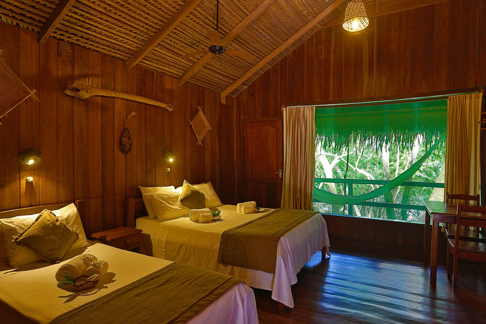 Intérieur d'un bungalow rustique au Juma Lodge, tout en bois avec un toit en paille. Deux lits simples avec des draps blancs et des jetés verts sont visibles. Des lampes murales en bois et une suspension en osier éclairent la pièce. Une grande fenêtre donne sur la forêt amazonienne et un hamac est suspendu sur le balcon. Une table et des chaises se trouvent près de la fenêtre.