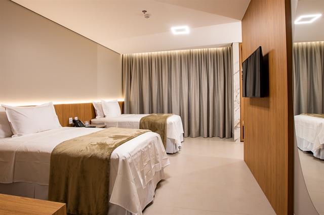 Intérieur d'une chambre d'hôtel triple avec trois lits simples. Les lits ont des draps blancs et des têtes de lit en bois avec éclairage LED. Il y a un bureau, un téléviseur mural et une grande fenêtre avec des rideaux.