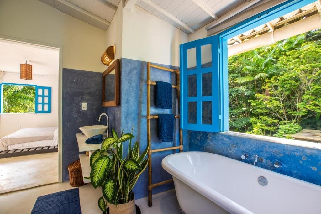 Vue d'une salle de bain à la Casa Ryokan avec une baignoire blanche et une grande fenêtre ouverte sur une forêt tropicale dense, encadrée de volets bleu vif et de murs texturés bleus. Il y a aussi un porte-serviettes en bambou avec des serviettes bleues, un évier et une plante en pot. Une chambre est visible en arrière-plan.