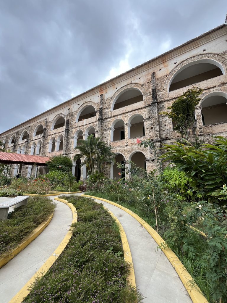 Vue en contre-plongée sur un grand édifice historique en pierre aux arcades superposées au Monastère des Jésuites à Baturité, précédé d'un jardin avec des parterres de fleurs et des chemins bordés de jaune.