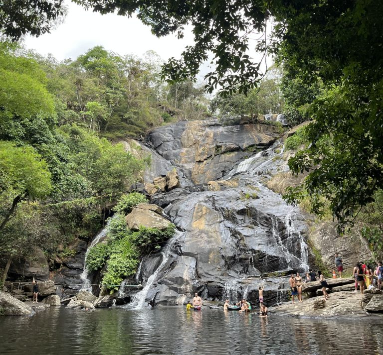 Grande cascade coulant sur des rochers sombres entourés d'arbres verts, avec un bassin naturel au premier plan où des gens se baignent et se détendent au pied de la chute d'eau à Guaramiranga.