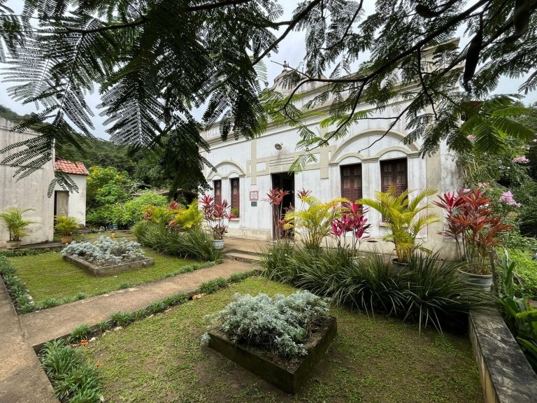 Maison coloniale centenaire blanche avec portes et fenêtres en bois à Mulungu, précédée d'un jardin soigné avec des parterres de feuillages colorés et ombragée par des branches d'arbres.
