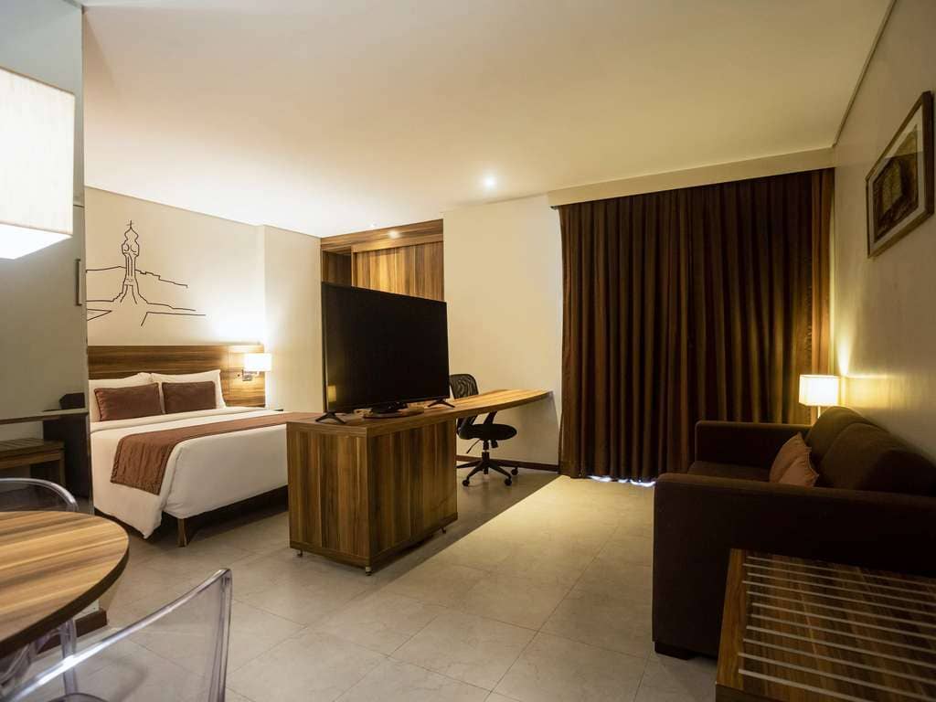 Une chambre d'hôtel moderne et bien éclairée au Mercure Belém Boulevard avec un grand lit confortable, un bureau avec une chaise, une télévision à écran plat, un canapé-lit confortable et une grande fenêtre avec des rideaux sombres. L'éclairage est chaud et l'atmosphère est accueillante.