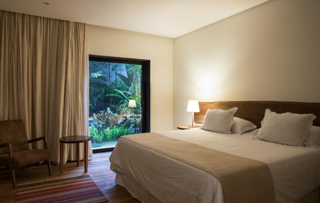 Chambre d'hôtel moderne et lumineuse avec lit king-size, mobilier en bois brut, fauteuil en cuir et grande baie vitrée s'ouvrant sur un jardin tropical luxuriant.