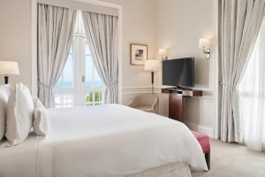 Une chambre élégante et luxueuse d'hôtel, dotée d'un lit king-size habillé de linge blanc, de rideaux gris clair, de murs aux tons neutres et de grandes portes-fenêtres offrant une vue panoramique sur l'océan à Rio de Janeiro.