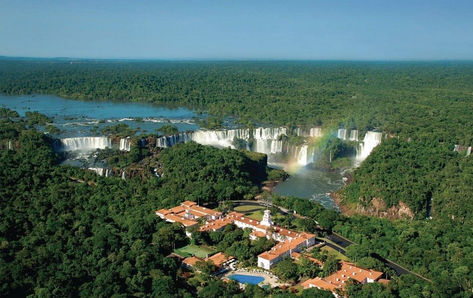 http://Vue%20aérienne%20des%20chutes%20d'Iguaçu%20entourées%20d'une%20forêt%20tropicale%20dense,%20avec%20un%20complexe%20hôtelier%20de%20luxe%20au%20premier%20plan%20et%20un%20arc-en-ciel%20visible%20dans%20la%20brume%20des%20cascades.