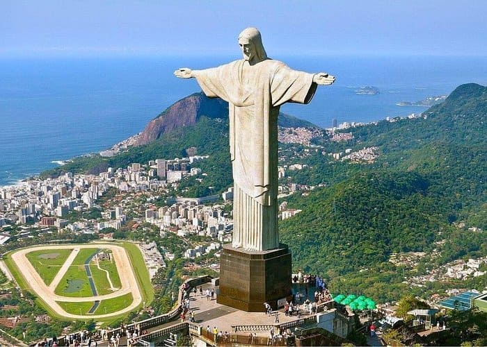 Vue aérienne de la statue monumentale du Christ Rédempteur aux bras ouverts, dominant la ville de Rio de Janeiro, les collines verdoyantes, un hippodrome et l'océan sous un ciel bleu dégagé.