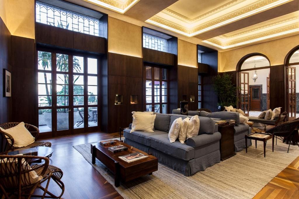Vue du spacieux hall d'entrée de l'hôtel Fasano à Salvador de Bahia avec des canapés bleus, des sièges en rotin, des boiseries sombres et de grandes portes vitrées lumineuses.